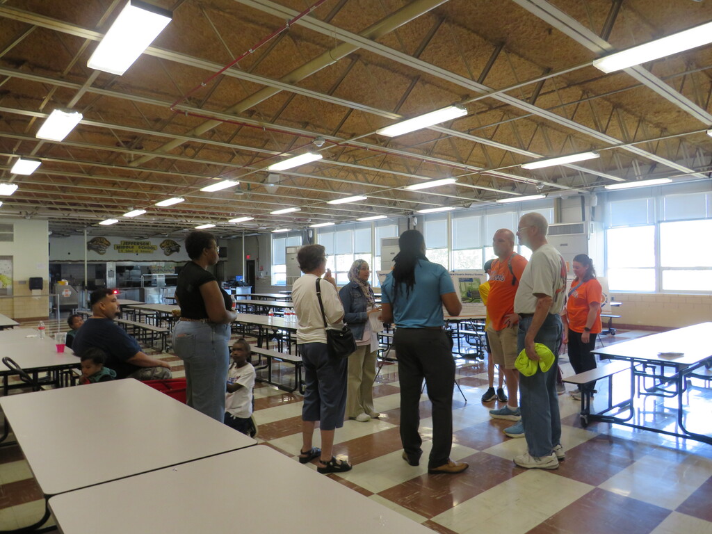 People stand around tables in a cafeteria. Long tables line the room, and overhead lights provide illumination.