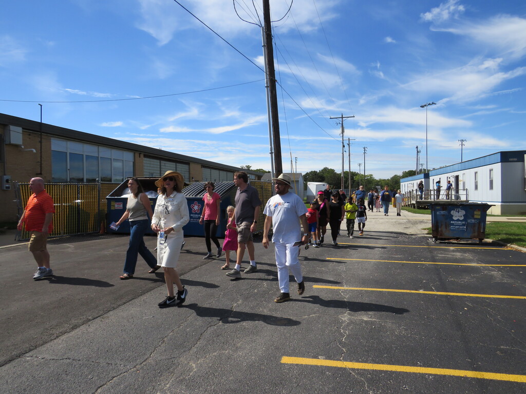 A group of people walks in a parking lot. Buildings, utility poles, and trash bins are in the background.