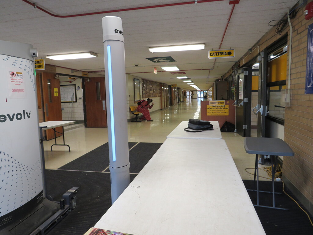 An empty corridor with white walls and floors. There are desks along the right side and a long table in the foreground.
