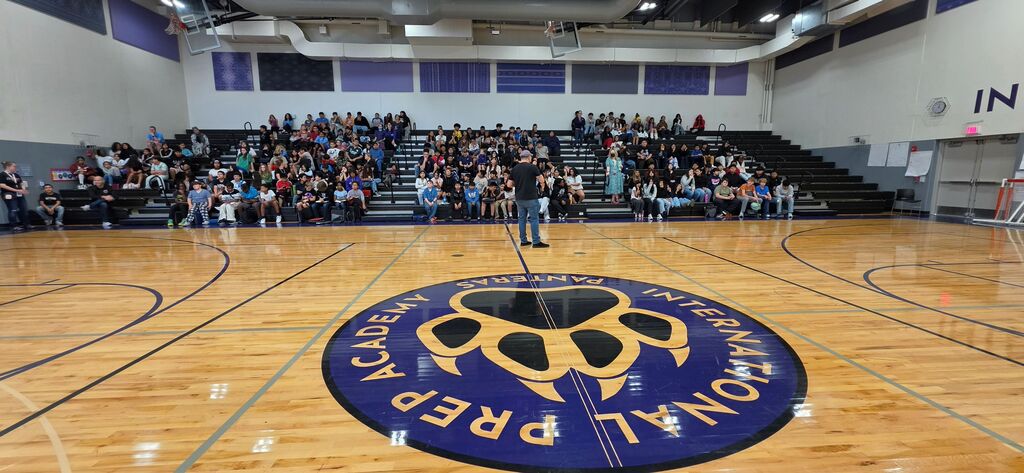 Interior of a gym with students seated on bleachers. A logo in the center has "International Prep Academy" written in purple.