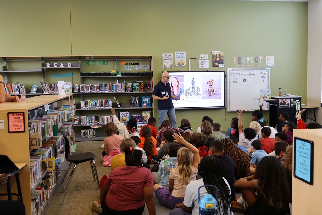 An author stands at a screen in a library. Many children sit on the floor listening to the author read.