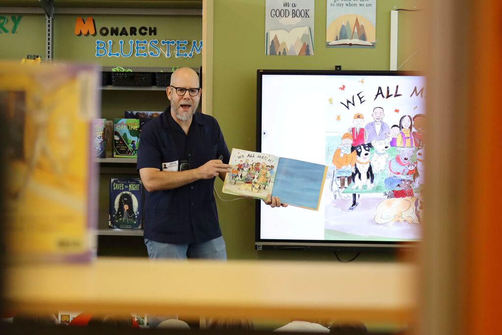 An author wearing glasses holds a book in front of a large screen projector. Behind them, a bookshelf displays books.
