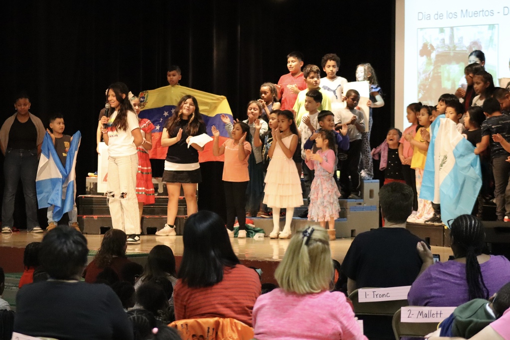 Children on stage holding flags of different countries, a projection screen behind them. Audience seated in front of the stage.