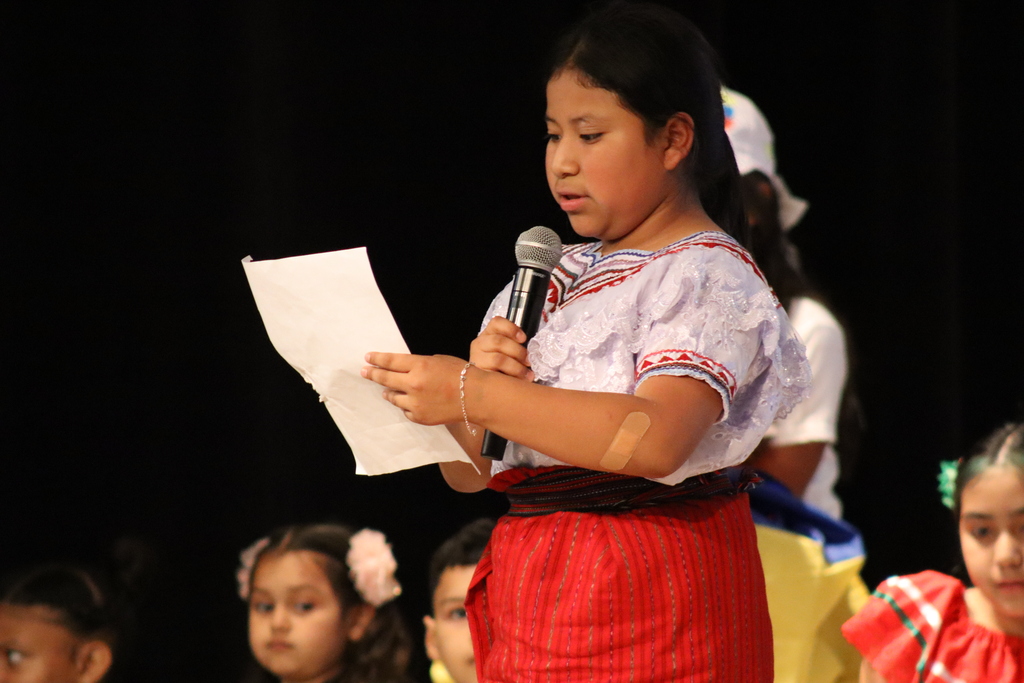 A young girl reads from a paper while holding a microphone, standing on a stage. Other children, some wearing colorful dresses, sit behind her.