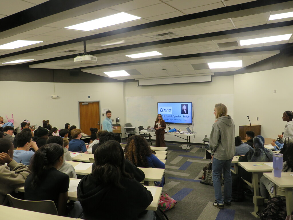 A classroom with students seated. A woman is standing in front of a screen and a person is standing near the front.
