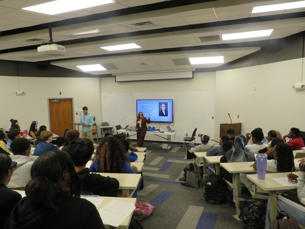 A classroom with desks and chairs, projector screen with a face, person standing in front, and students sitting.