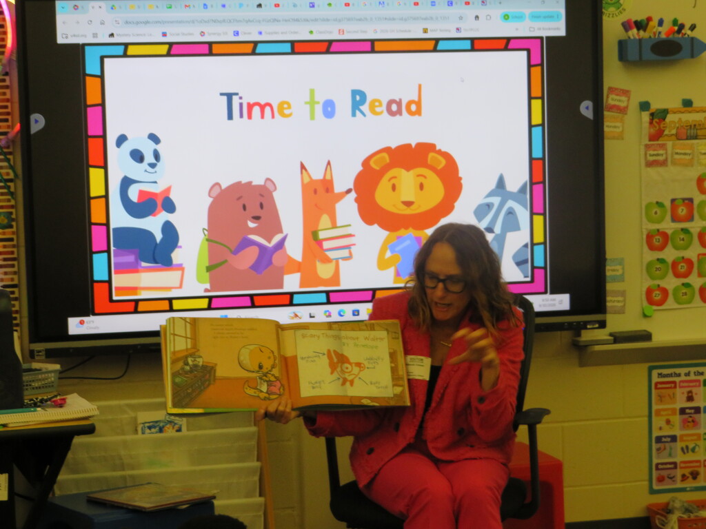 Woman in red outfit reading book to class, large screen behind displaying "Time to Read" with animal characters.