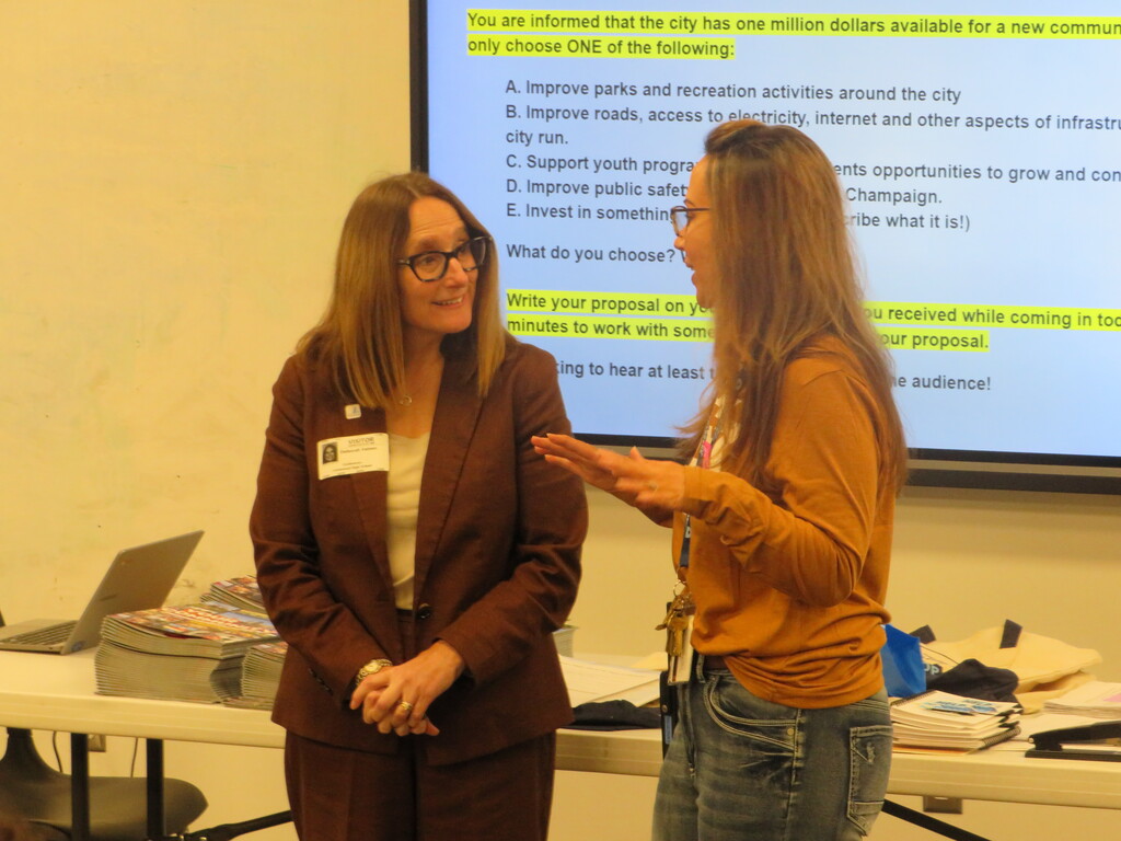 Two women talk in front of a screen with a city budget proposal. One wears glasses and a badge.