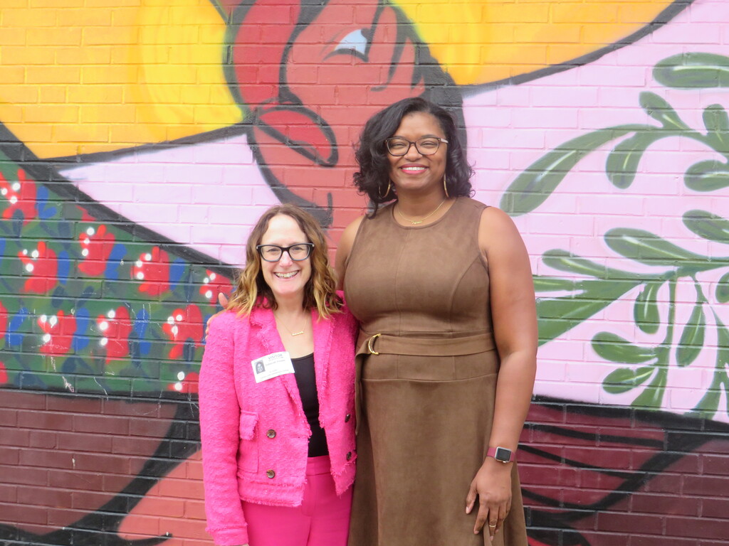 Two women in front of a wall with multicolored artwork. One wears a pink blazer, the other a brown dress.