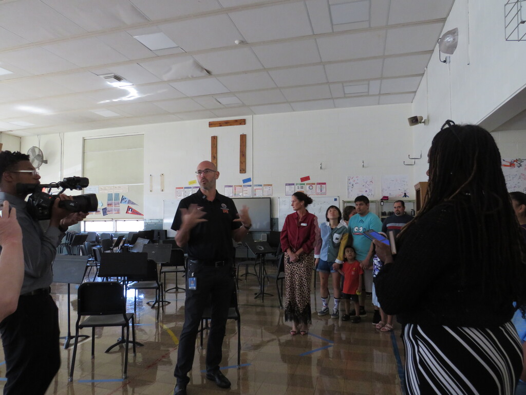 Group in a room, one person speaking, another holding a camera. Musical instruments on floor.