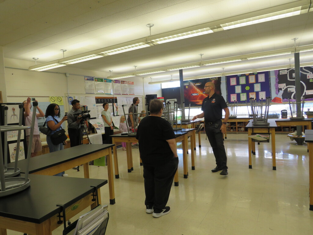 Indoors, a classroom with tables. A man stands with students, speaking. A group of people stands near the wall.