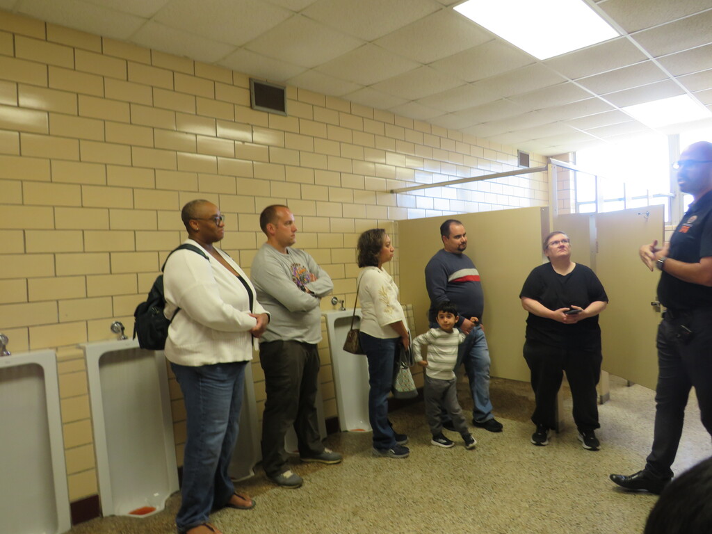 A group of six people stands in a public bathroom, with tiled walls and a speaker on the wall.
