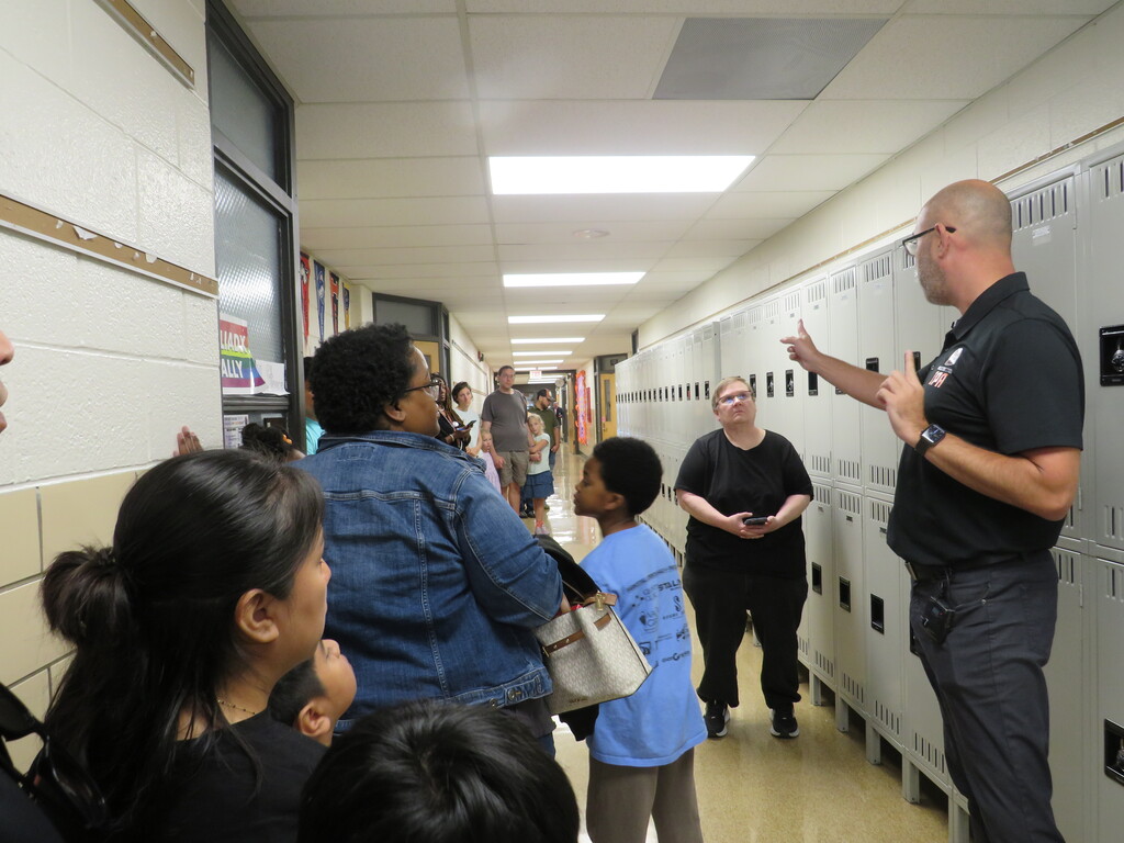 Several adults and children are in a school hallway. One adult points at a locker while others watch.