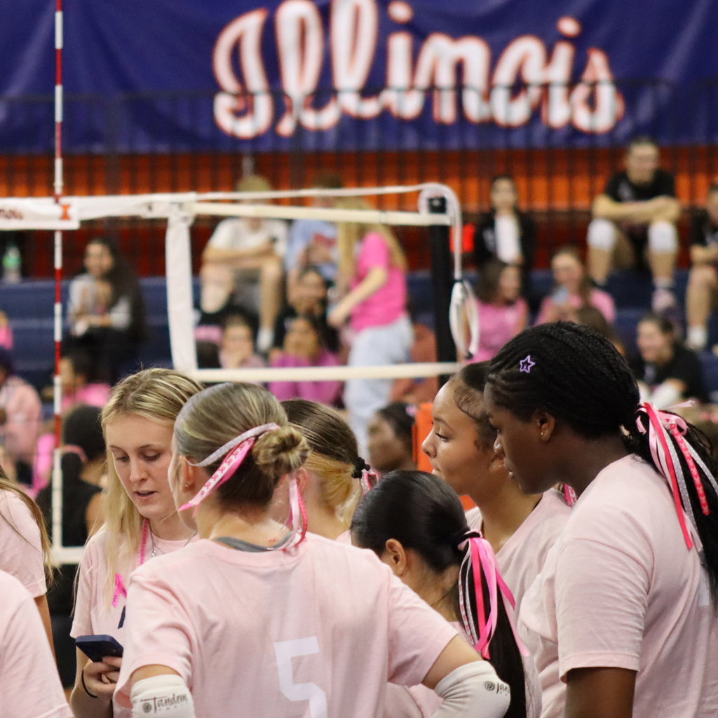 Centennial High School volleyball players take a time out during game at Huff Hall. The word "Illinois" in orange and blue can be seen in the background.