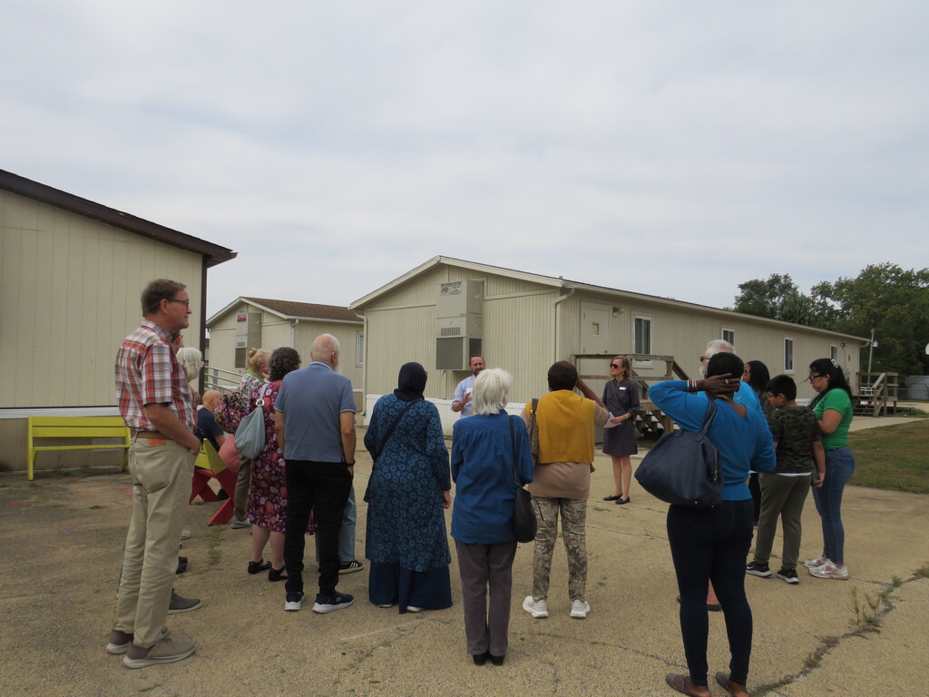 A group of people stands in a circle outside near buildings. Some carry bags. A yellow bench is visible.
