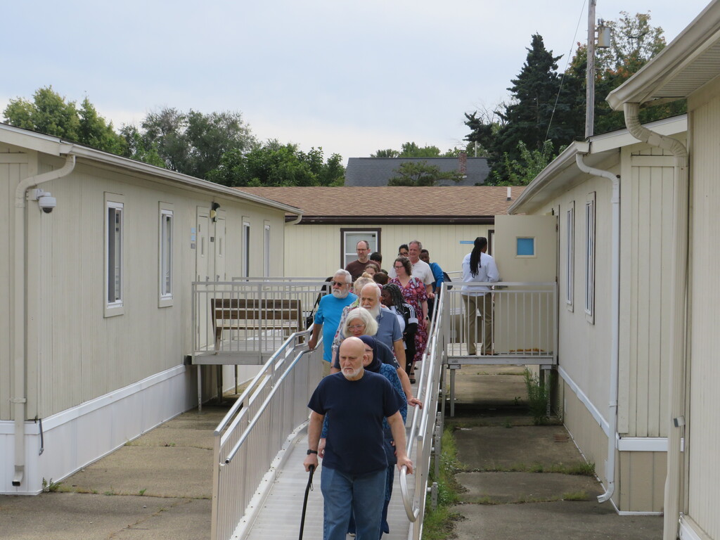 People walk on a pathway between mobile homes. Some use canes. Trees and a building are visible in the distance.