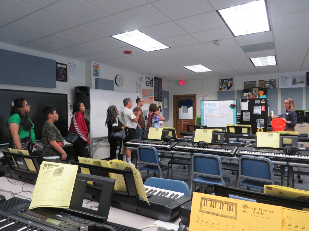 A music classroom with musical instruments. People are gathered around a teacher at the front of the room.