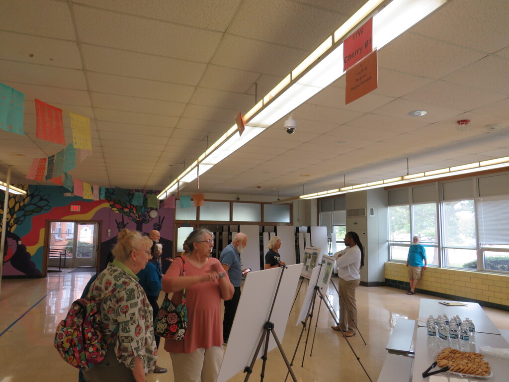 People view an art exhibit in a large room. A mural decorates the wall. Bright lights illuminate the area.