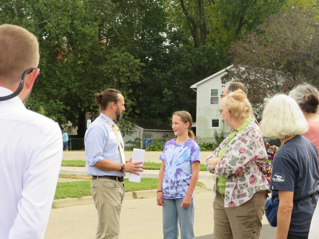 A group of people standing on a street. One holds a paper, and they are surrounded by green trees and a house.