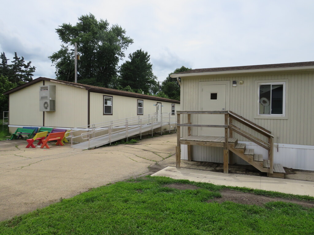 Two modular homes, one with a ramp, set against a backdrop of trees. The front yard has grass and a walkway.