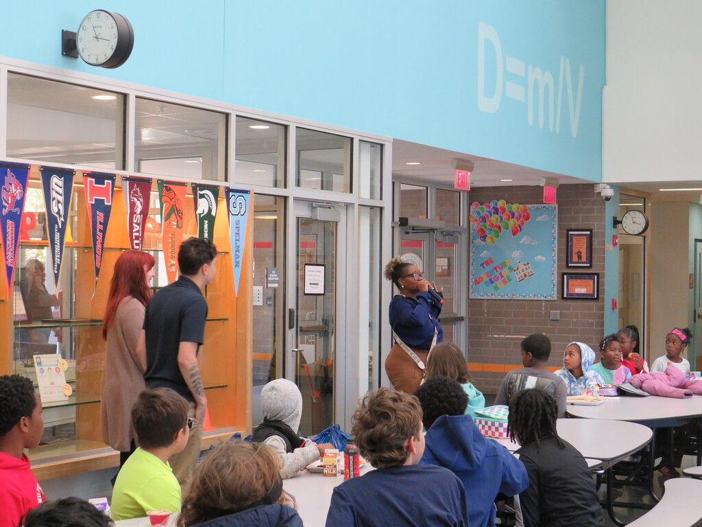 Hallway with glass walls, a clock, and people standing and sitting around tables.