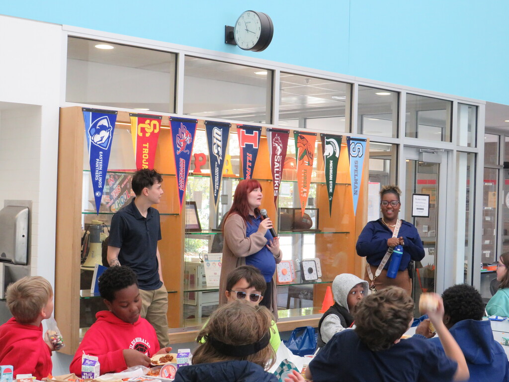 People inside a cafeteria. Some are standing, and some are sitting. A woman holds a microphone.