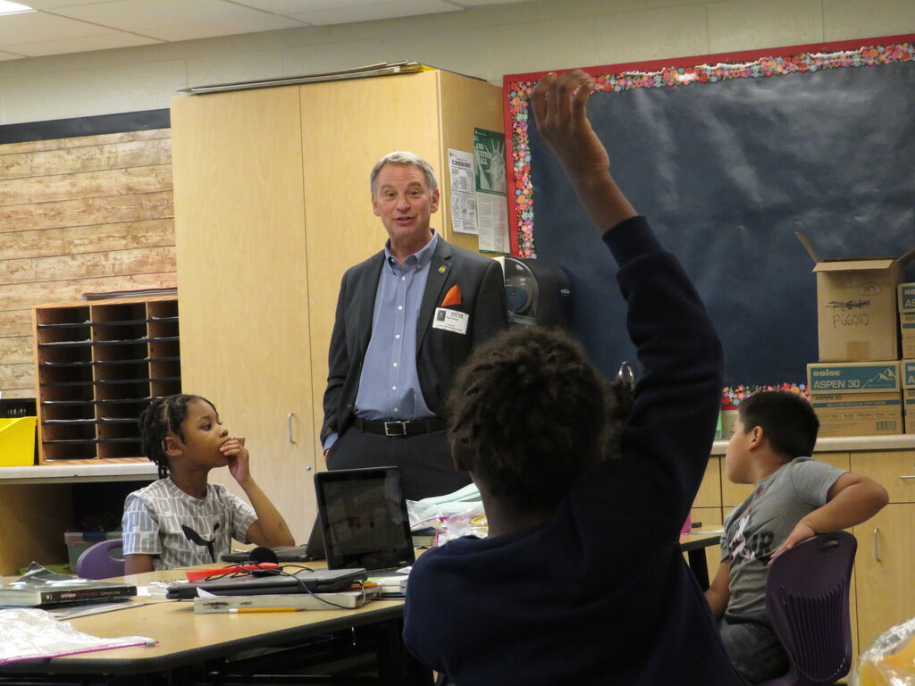 A classroom with a teacher standing near a desk with a laptop. Students sit at desks, one raising a hand.
