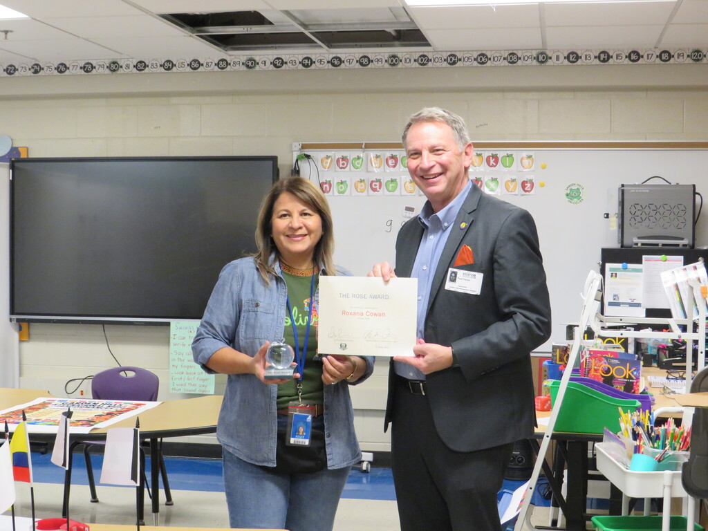 A woman and a man holding a framed certificate stand in a classroom with chairs, tables, and a whiteboard.