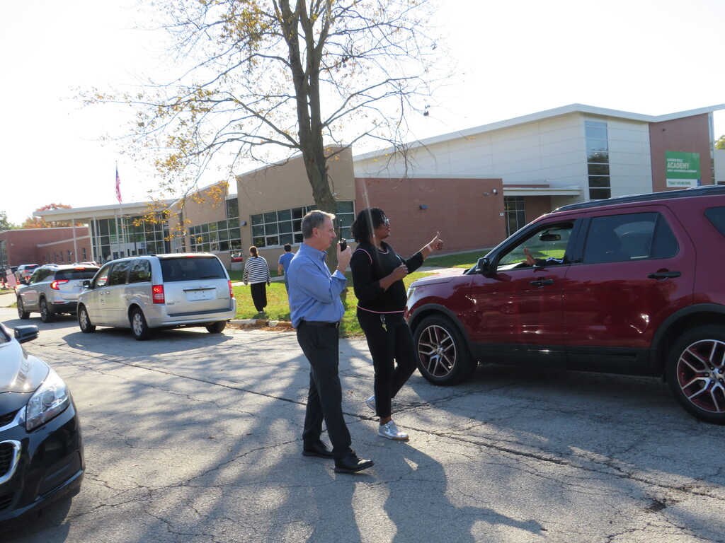 A man speaks into a microphone, gesturing with his hand, next to a woman by a red SUV.