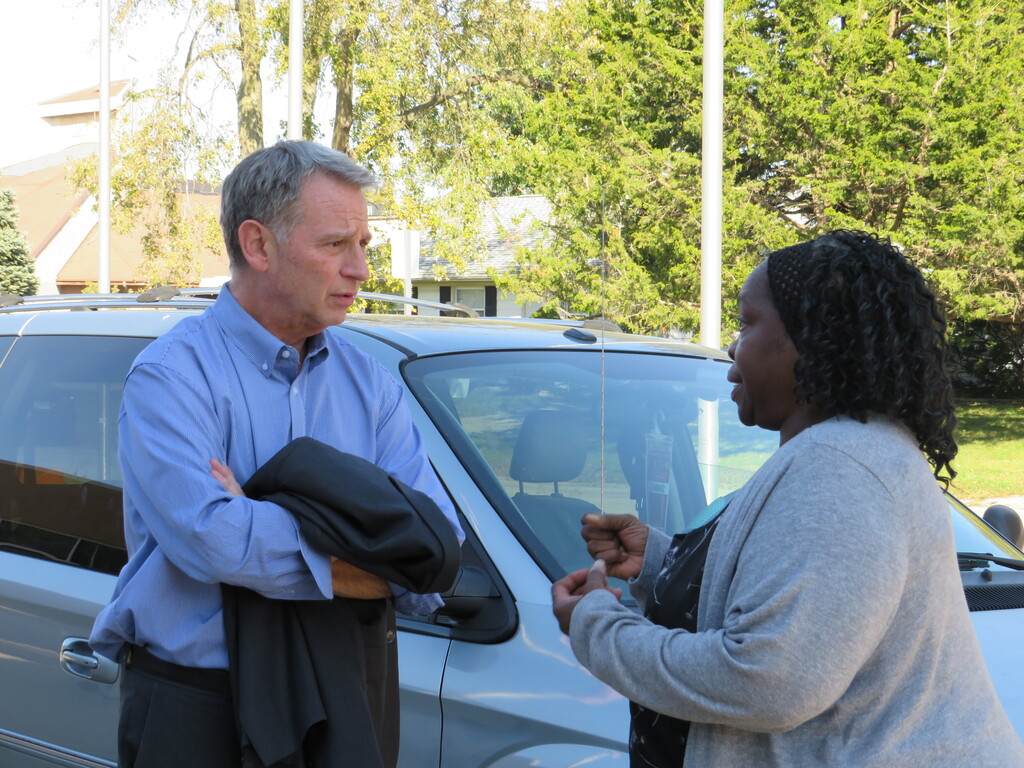 A man and a woman standing near a car, man holding a jacket and speaking to the woman.