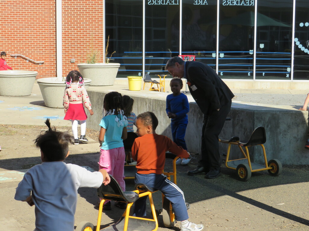 Man in suit bends to speak to children on small bikes and in carts in a courtyard.