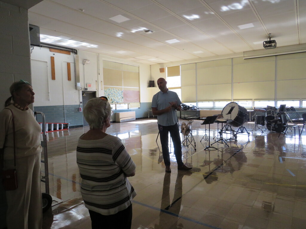 Three people in a large room with musical instruments on the floor, a man stands and talks.