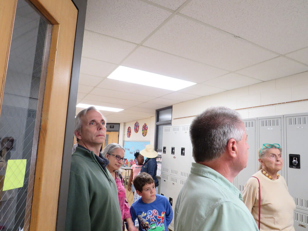 People stand in a hallway, some looking through glass doors. Lockers line a wall, and a yellow post-it is stuck to a door.