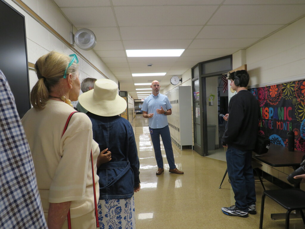 People stand in a hallway. A man speaks to others near an open door. Murals are on the wall.