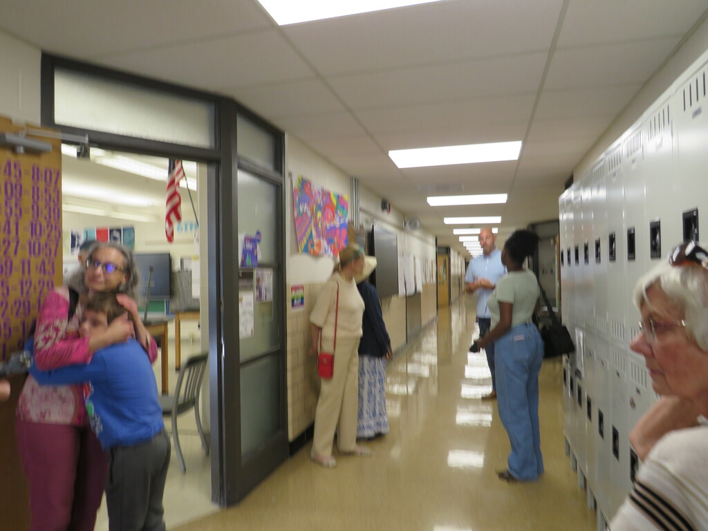 People standing in a hallway with lockers. A woman hugs another person. A flag is on the wall.