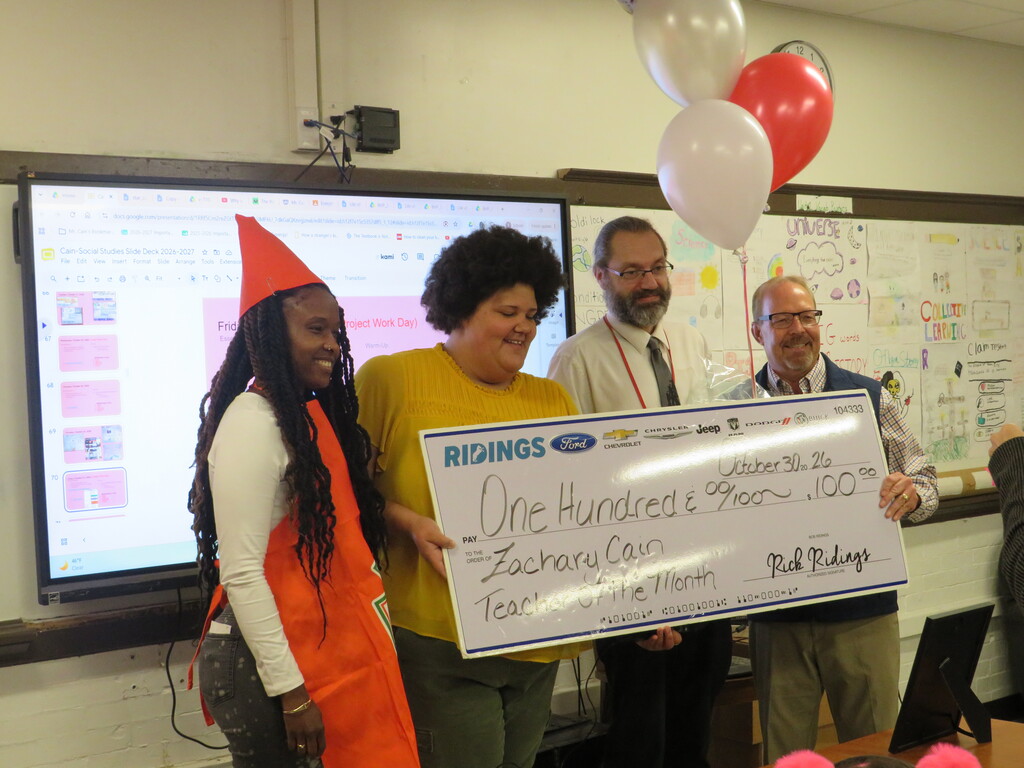 Five people stand behind a large check, smiling, while a whiteboard with a diagram is in the background.