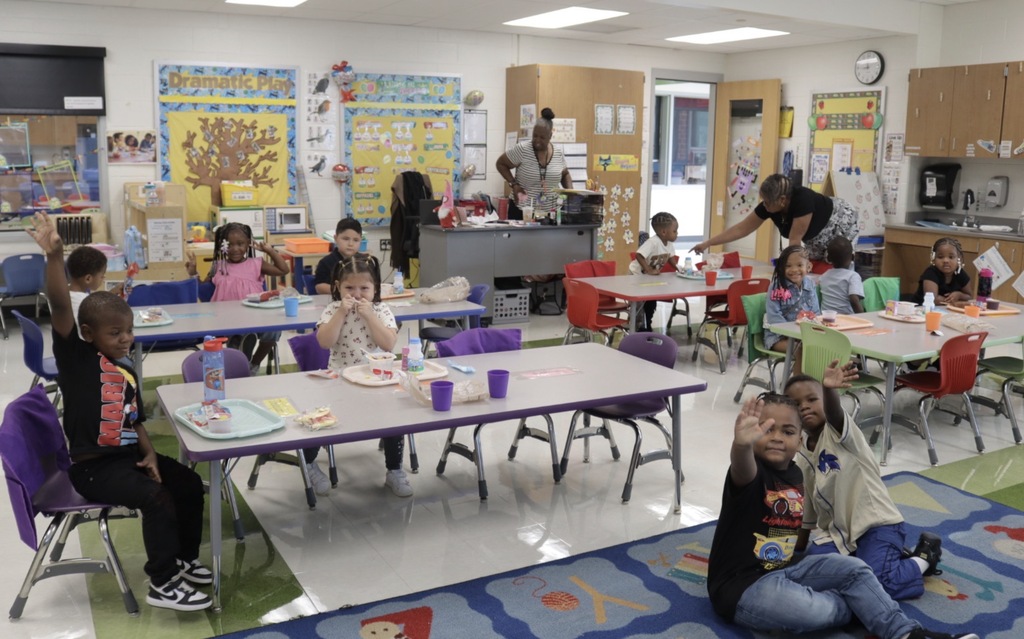 Pre-K students sit at classroom tables during their first day while teachers help them get settled.