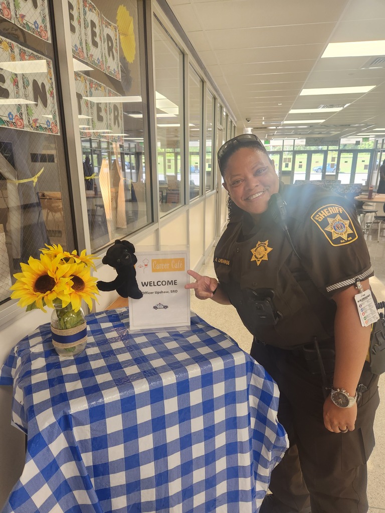 picture of school officer standing by a table with a checkered tablecloth, a vase of flowers and a sign