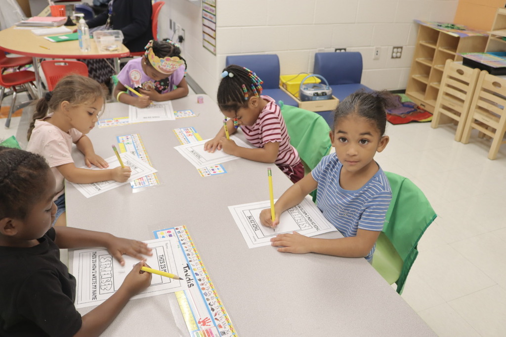 Pre-K students sit at a classroom table practicing writing and early literacy skills on worksheets.