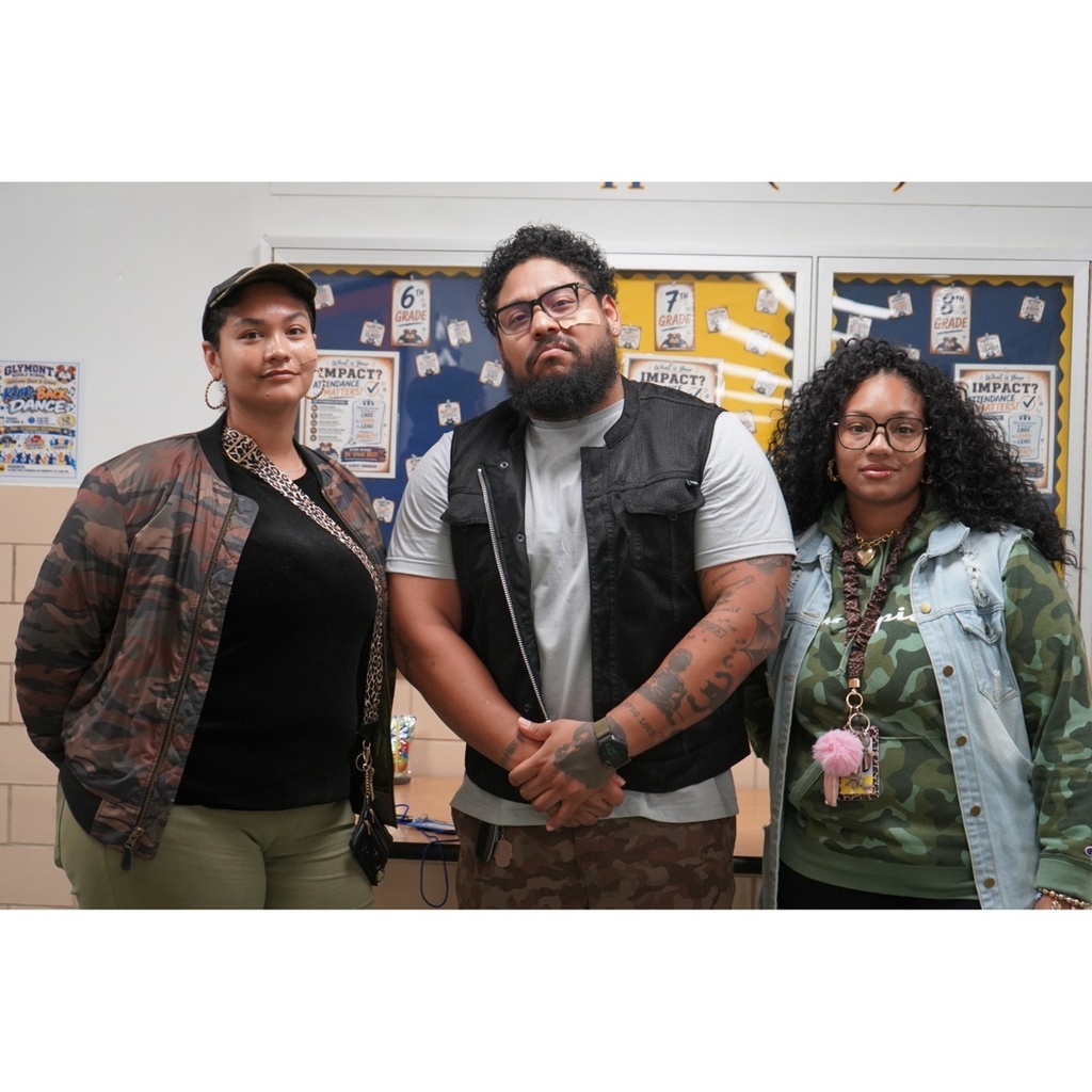 Three adults pose together indoors during a school Twin Day celebration. The two people on the left wear camouflage-patterned clothing, while the person in the center wears camouflage-patterned pants, creating a coordinated matching look. The group stands in front of school bulletin boards displaying grade-level information and attendance posters. The photo was taken in a brightly lit school hallway or common area as part of Unity Week activities.