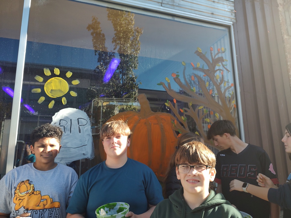 middle school students pose with painted window with sun, gravestone , pumpkin, and fall tree