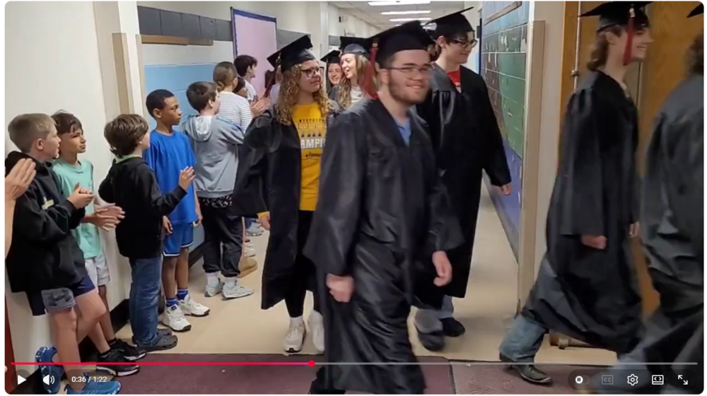Seniors in graduation caps and gowns walking through MED Elementary School while students calp.