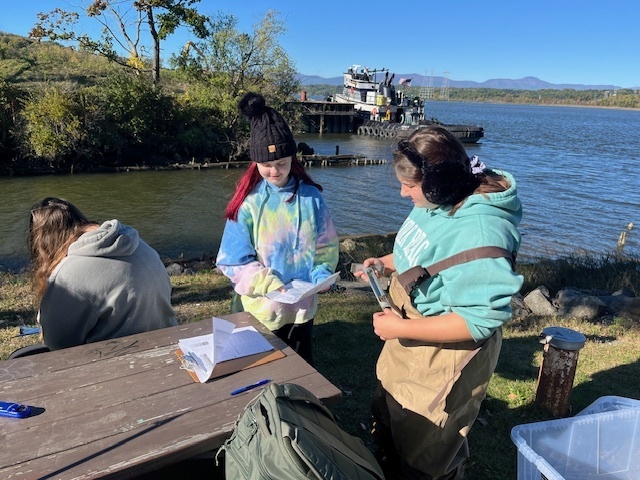 students logging data at picnic table alongside river 
