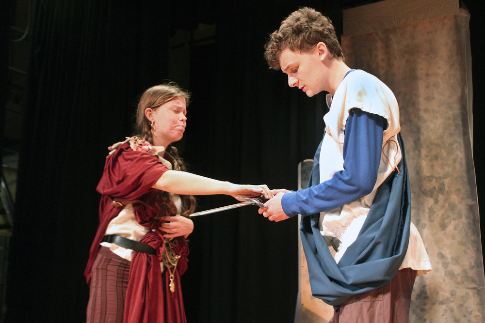 girl handing blade to boy on stage in roman dress