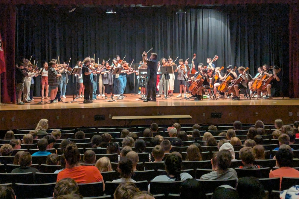Students and adults playing string instruments on stage before an audience of students