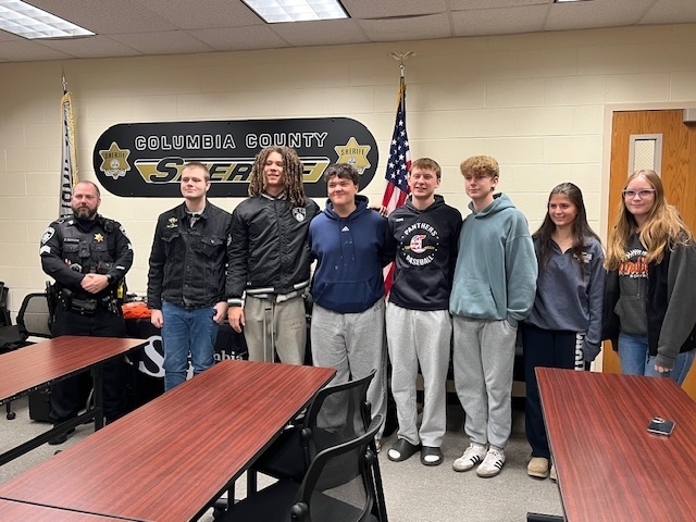 teenage students and sheriff's deputy pose for group photo in front of Columbia County Sheriff's sign
