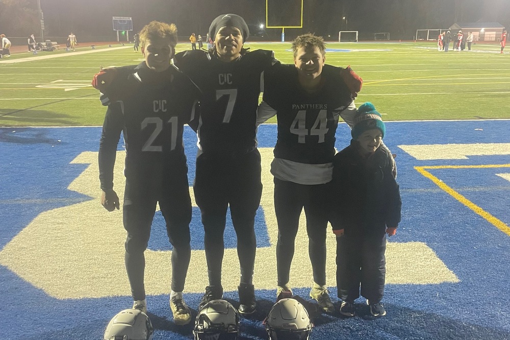 three high boys in CIC football uniforms  pose with young boy on football field.