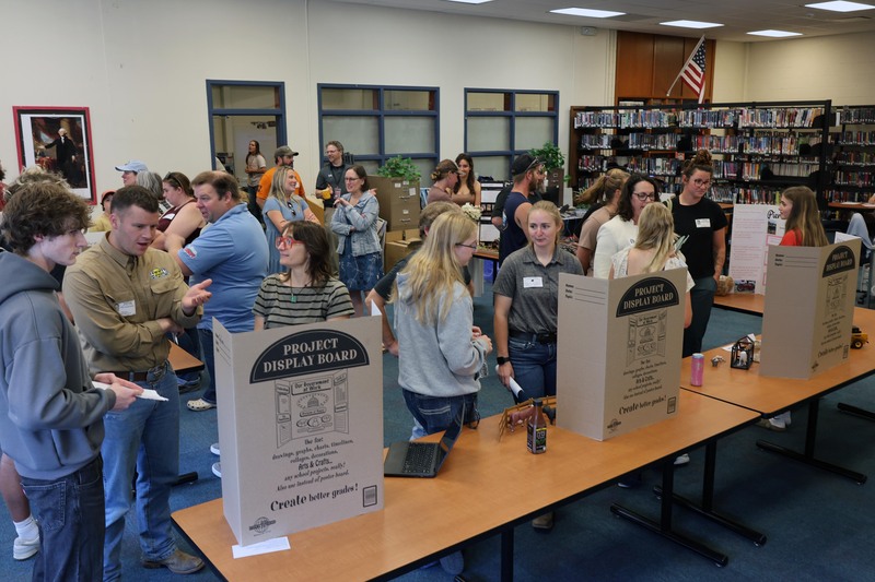 large group of adults and students look at display boards set up in high school library