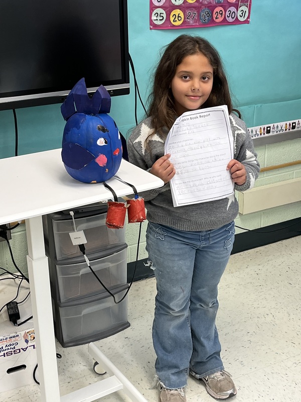 girl holding book report next to decorated pumkin
