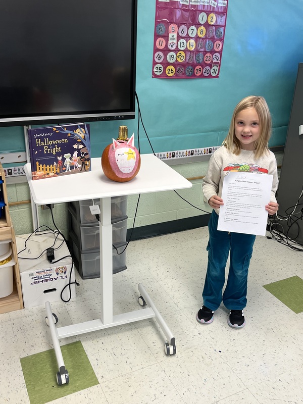 girl holding book report next to book and decorated pumpkin